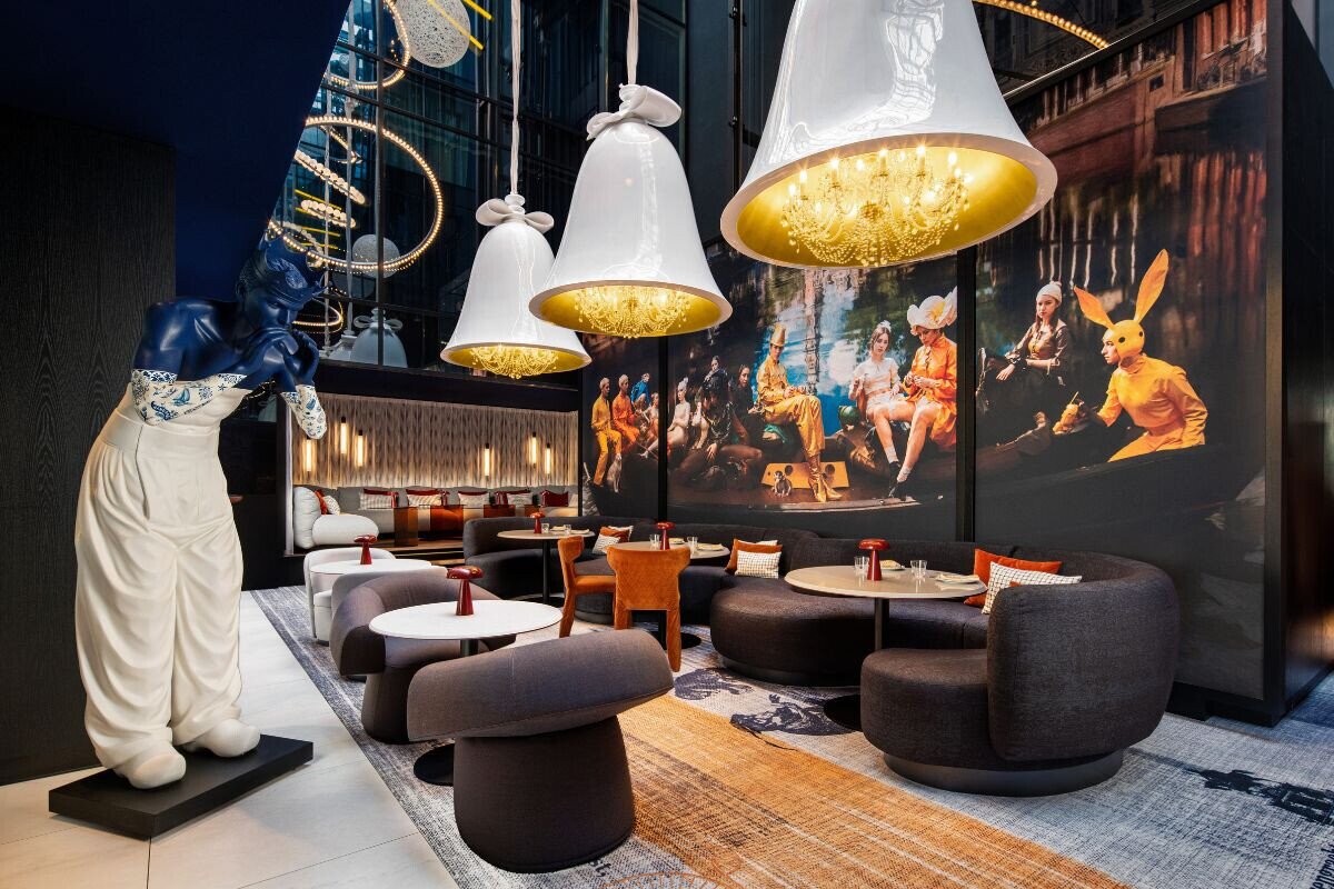 The Dutch design interior at Bar Restaurant Prins & Aap featuring bell-shaped chandeliers with golden interiors, plush curved seating, and a striking mural triptych of people in theatrical costumes. The blue Marcel Wanders Prince statue creates a sophisticated yet playful ambiance.